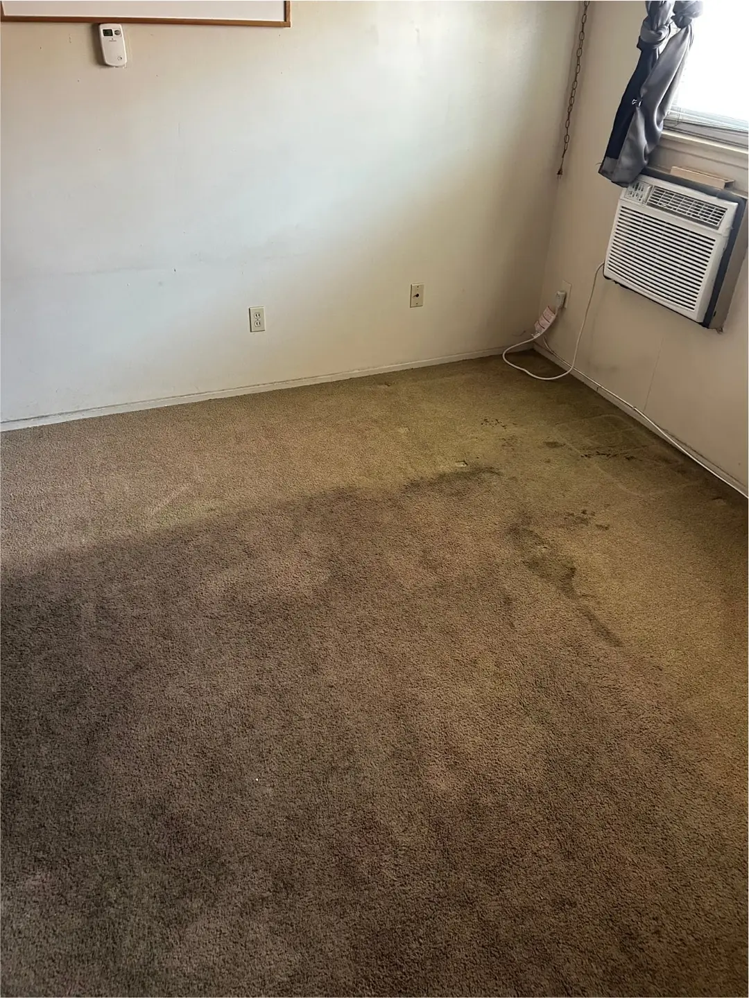 Carpeted room with dark soil marks near a wall and window air conditioner before cleaning